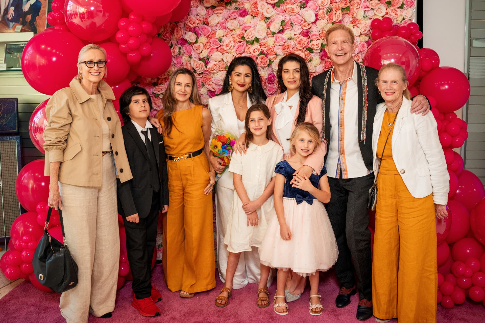 Multi-generational group of guests posing on the pink carpet in front of a rose wall and pink balloon garland
