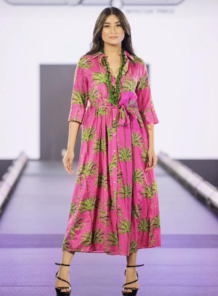 Model in a pink palm-print shirt dress with a green link necklace posing on the runway