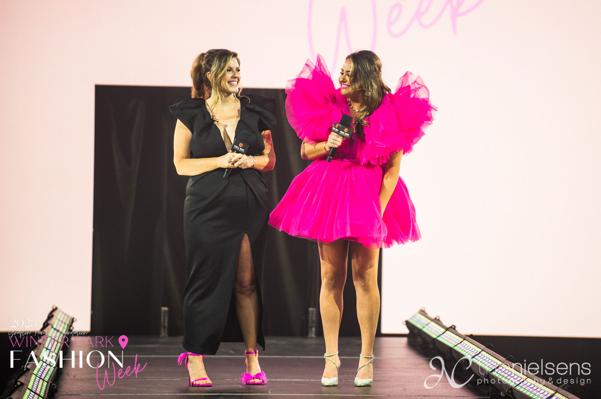 Two hosts in black and hot-pink gowns presenting the show from the runway