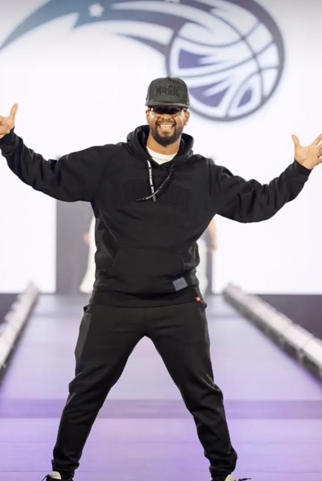 Smiling model in a black hoodie, joggers and cap posing with arms wide on the runway